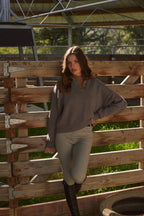 Woman in a brown sweater and beige pants standing in front of a wooden fence with greenery in the background.
